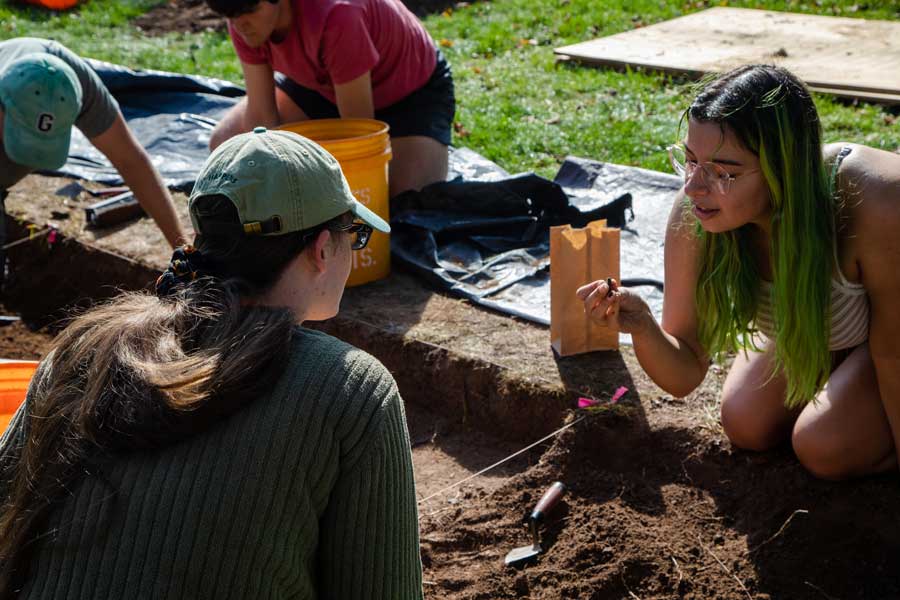 Students sitting on the ground in front of an archeological dig site